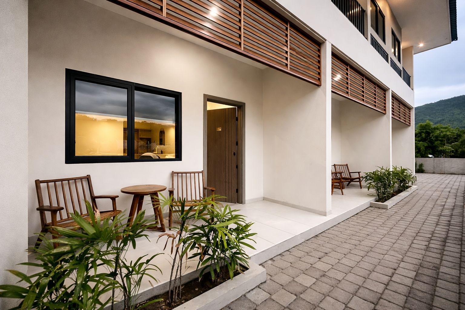 A modern building with a flat roof, a balcony with wooden railings, and a paved outdoor area with seating.