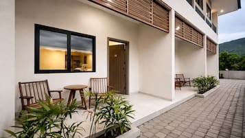 A modern building with a flat roof, a balcony with wooden railings, and a paved outdoor area with seating.