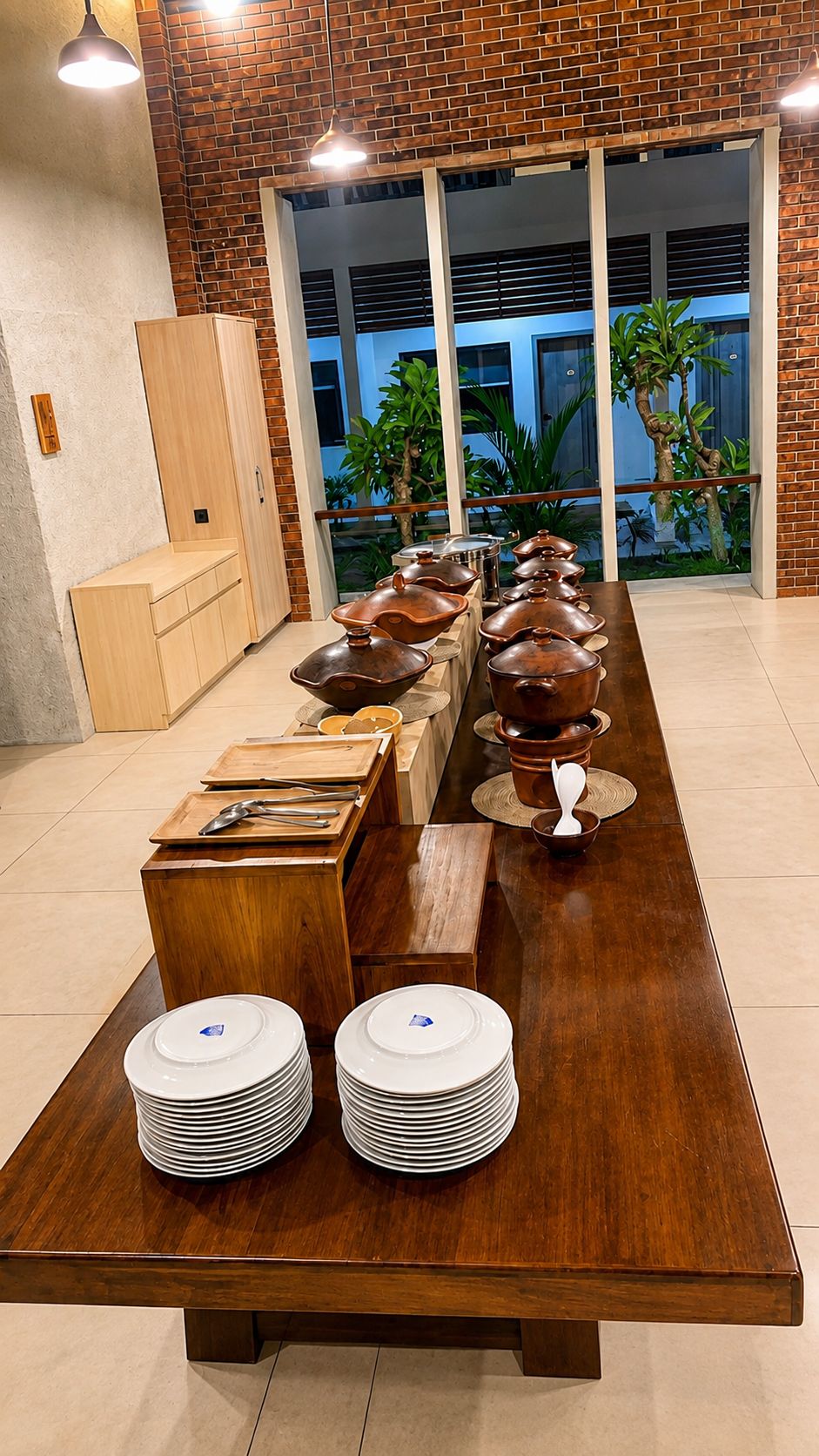 A long wooden table with stacked white plates and bowls, set against a brick wall with a view of greenery outside.