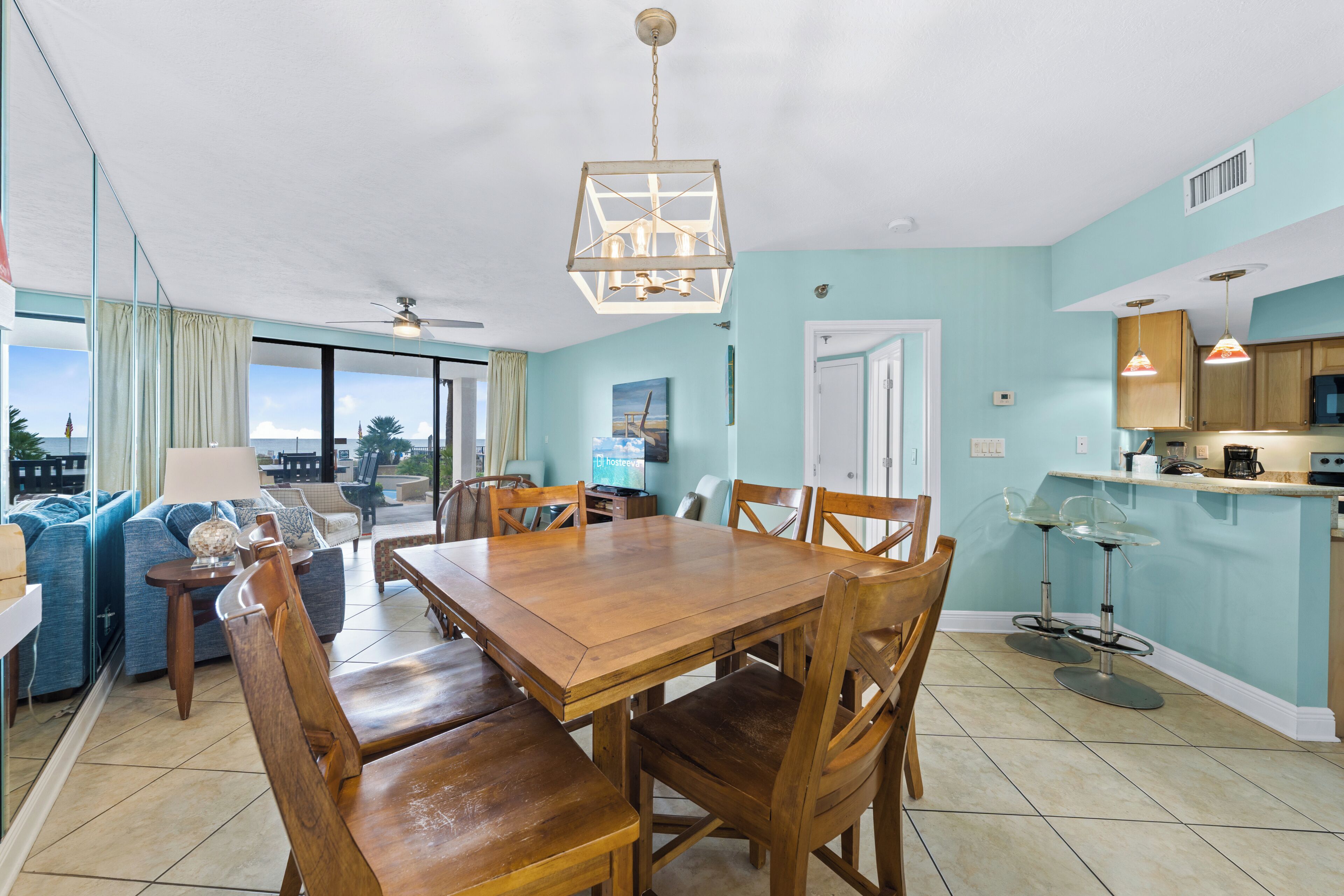 Een eetruimte met een houten tafel en stoelen, een keuken met blauwe wanden, en uitzicht op de oceaan door grote ramen.