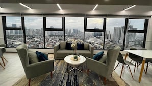 Ein moderner Arbeitsraum mit großem Fensterblick auf eine Stadtansicht, einem zentralen Sofa und einem kleinen runden Tisch mit Blumen.