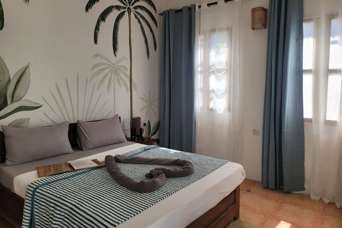 A bedroom with a bed, pillows, a striped blanket, a wooden headboard, a nightstand, a lamp, and a window with curtains.