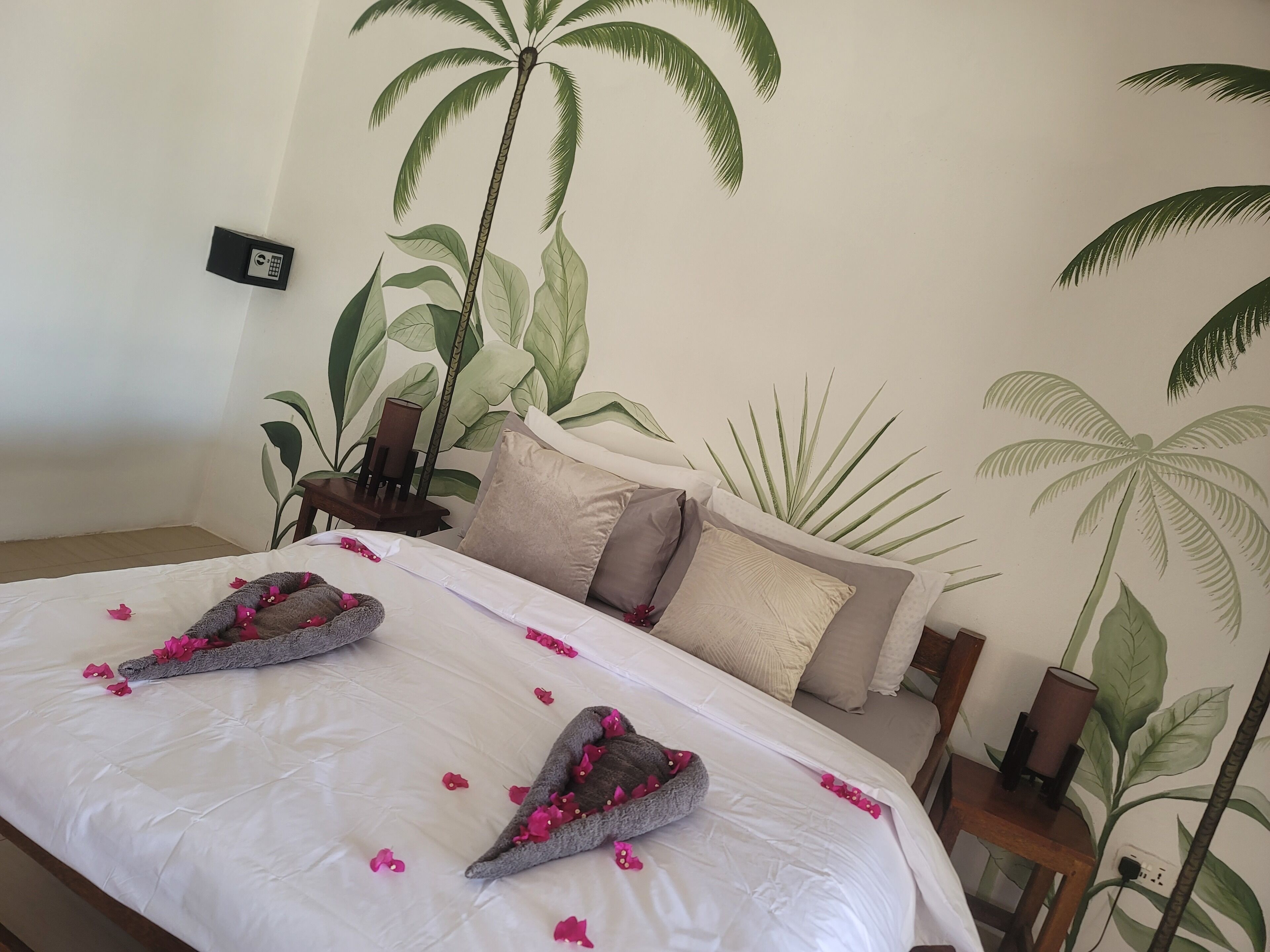 A hotel room with a bed, pillows, a nightstand, a lamp, and a wall mural of palm trees.