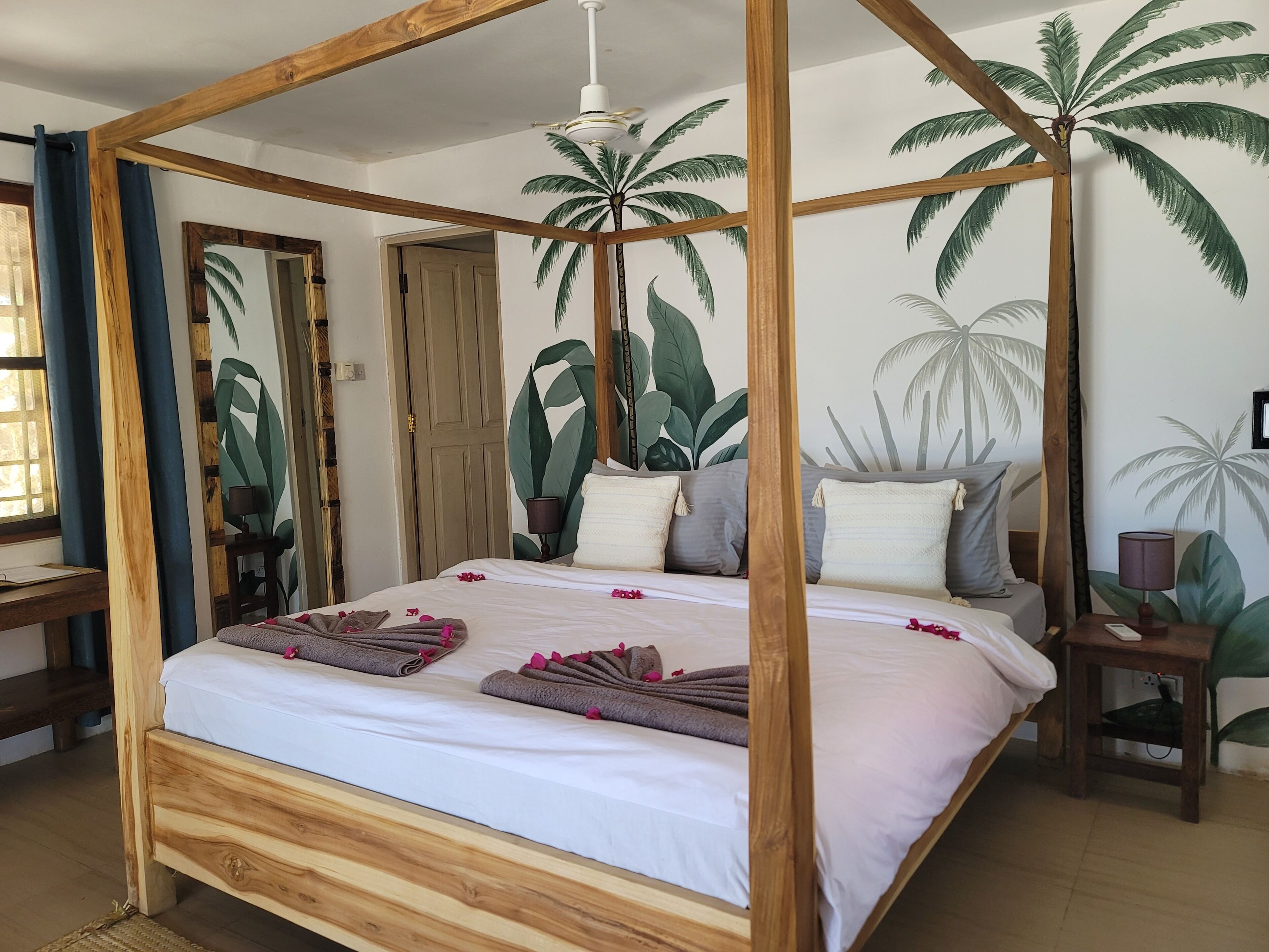 Ein hölzernes Himmelbett mit weißer Bettwäsche, ein Deckenventilator und ein Wandbild mit tropischen Pflanzen.
