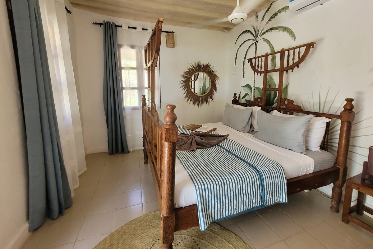 A bedroom with a wooden four-poster bed, a ceiling fan, an air conditioning unit, and a wall mural of palm trees.