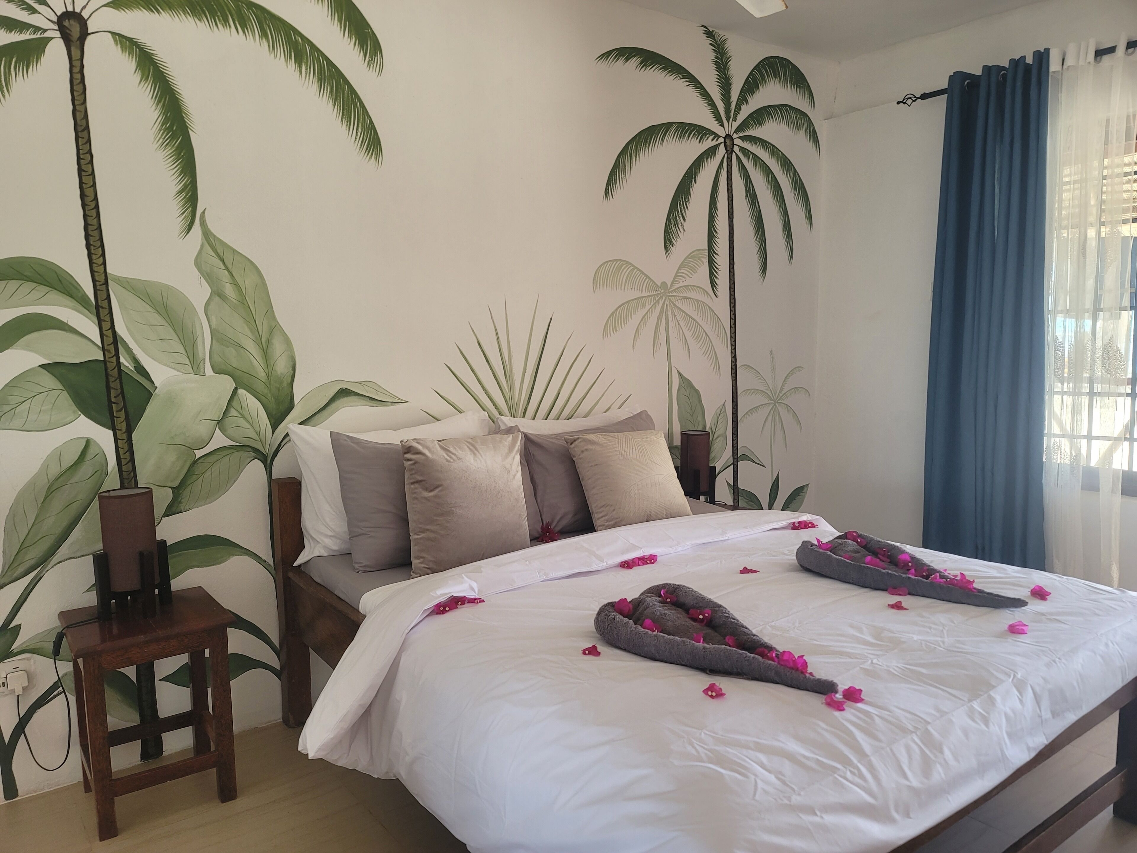 Un dormitorio con cama, almohadas, una mesita de noche, una silla de madera y un mural de palmeras.