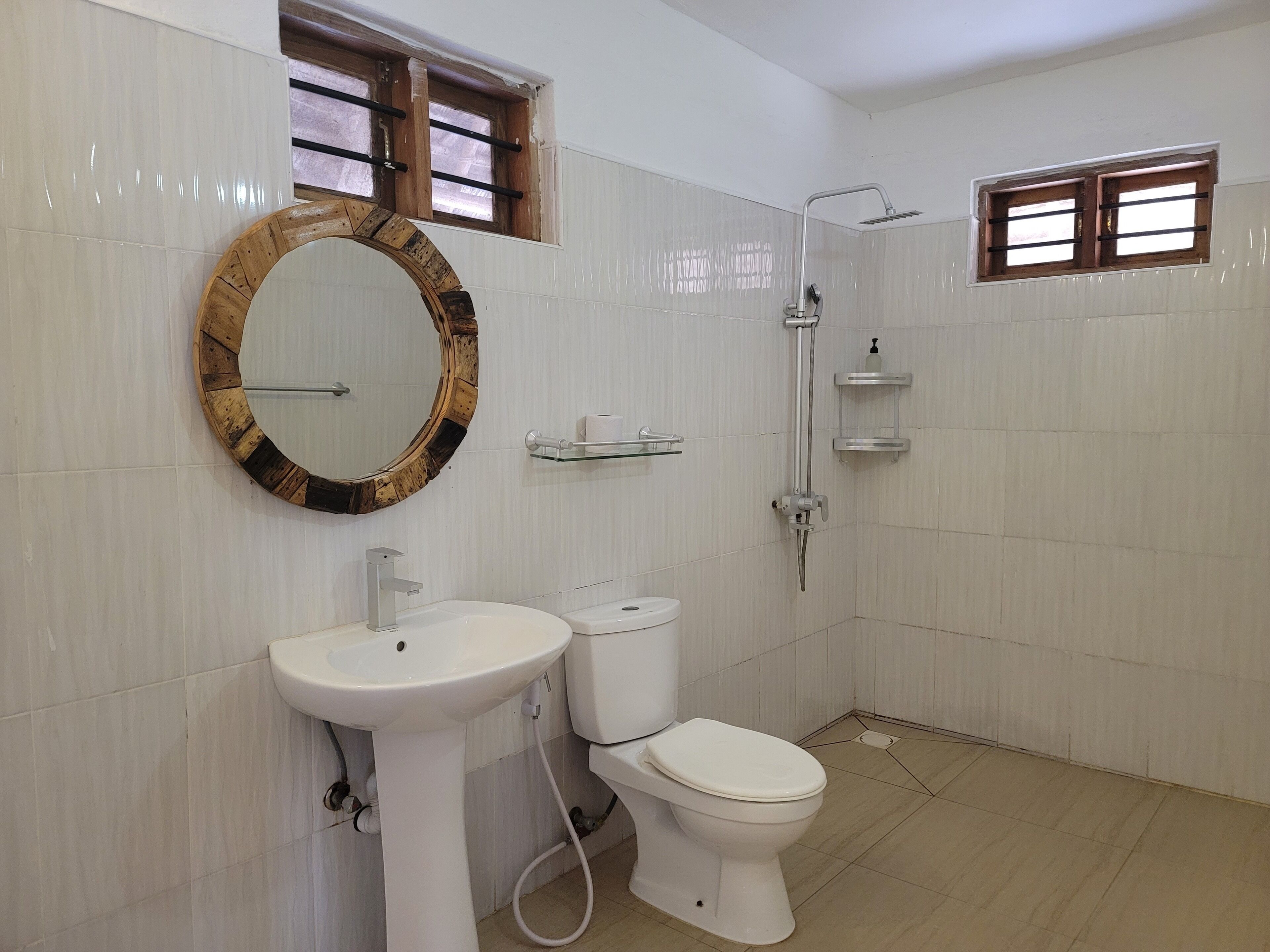 Une salle de bain avec un lavabo, des toilettes et une douche, le tout de couleur blanche. Un miroir à cadre en bois est placé au-dessus du lavabo, et il y a deux fenêtres avec des barreaux en bois.