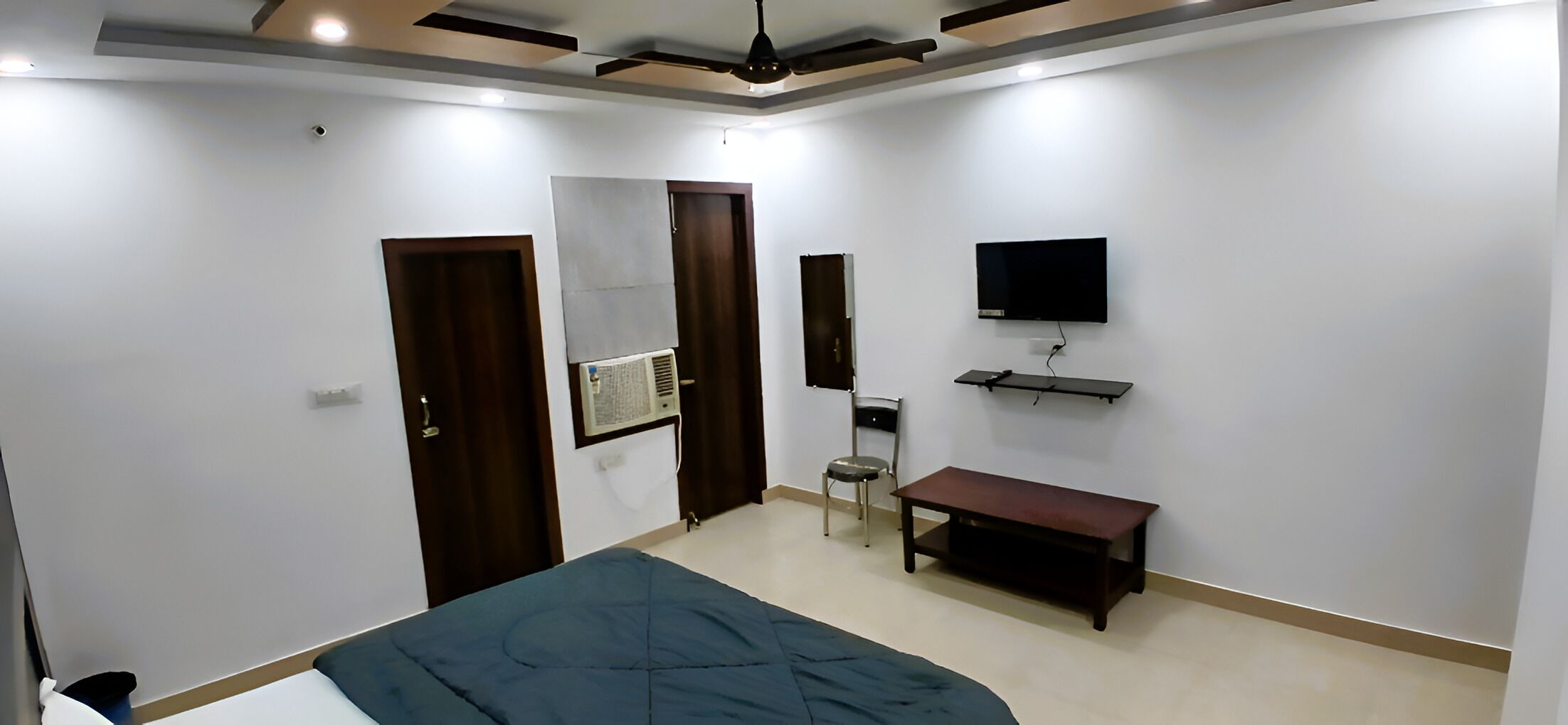 Un dormitorio con cama, una silla, un televisor montado en la pared y una mesita.