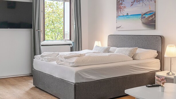A neatly made bed with white linens, a grey headboard, and a bedside table with a lamp and remote control. - Cozy 2BR Stay Near AKH With WiFi (Vienna)