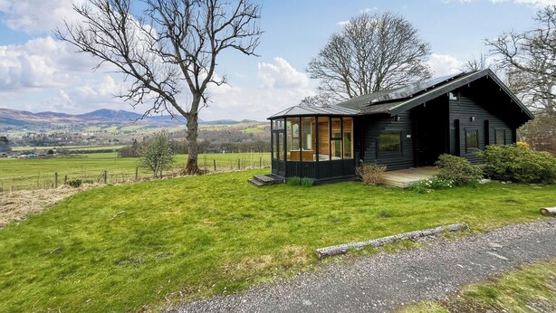 Een moderne zwarte chalet met een glazen uitbouw, gelegen in een grasveld met uitzicht op een berg.