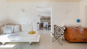 Amplia sala de estar con un sofá blanco, una silla estampada y un mueble de madera. Se ve una cocina al fondo.
