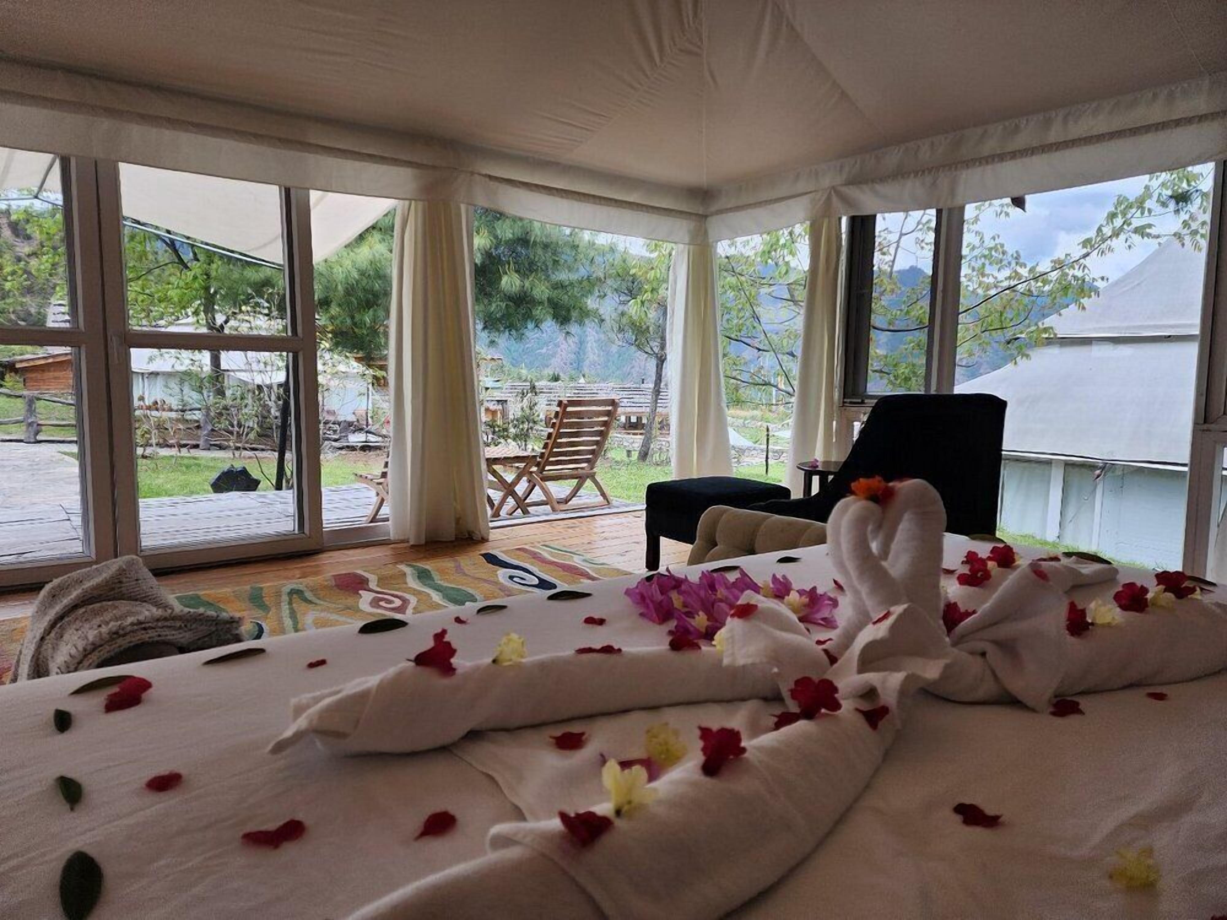 A room with a bed covered in white linens and rose petals, large windows, and a view of the outdoors.