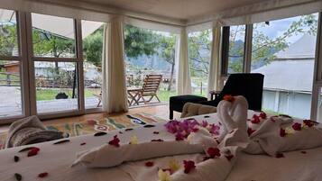 A room with a bed covered in white linens and rose petals, large windows, and a view of the outdoors.