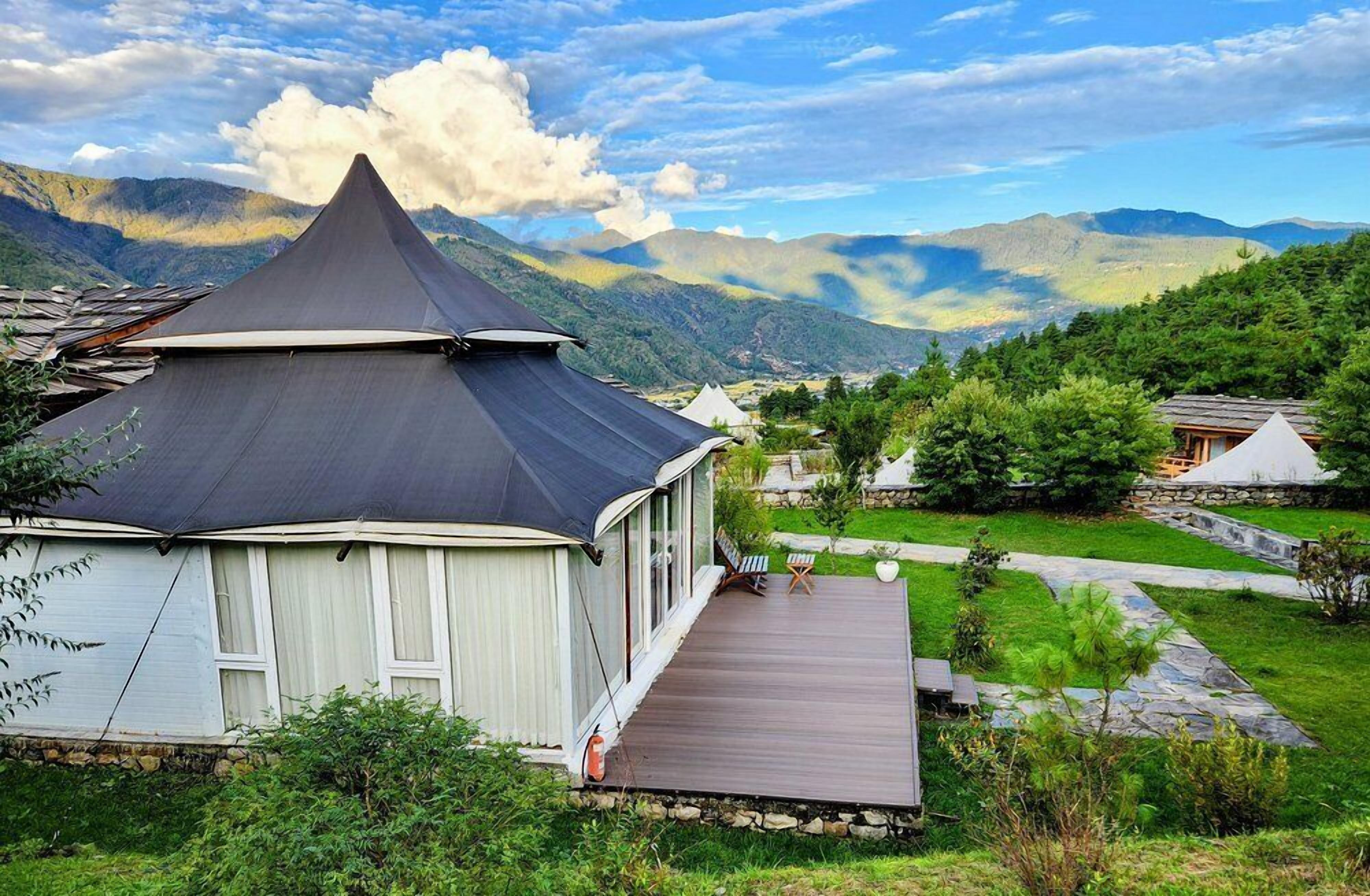 A traditional yurt with a conical roof, surrounded by a lush garden and a mountainous backdrop.