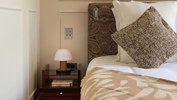 Een netjes ingerichte hotelkamer met een bed, een nachtkastje met een lamp, een wandlamp en een lijst met een kunstwerk boven het bed.