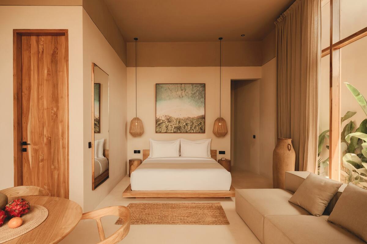 Ein modernes Schlafzimmer mit einem großen Bett, einer Couch, einem Esstisch mit Früchten und einem großen Fenster mit Vorhängen.