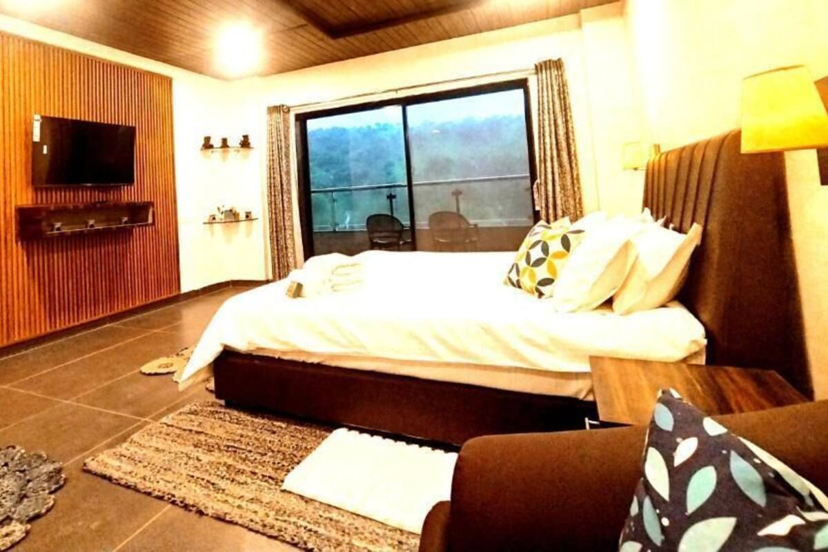 A hotel room with a large bed, a flat-screen TV mounted on a wooden panel, a balcony with a view, and a sofa.