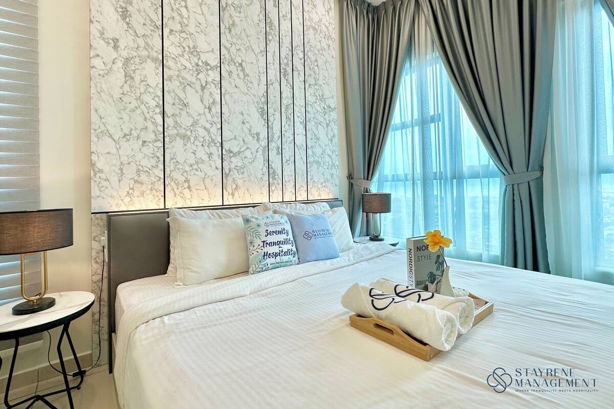 A hotel room with a marble-patterned wall, a neatly made bed with pillows, a bedside table with a lamp, and a window with curtains.