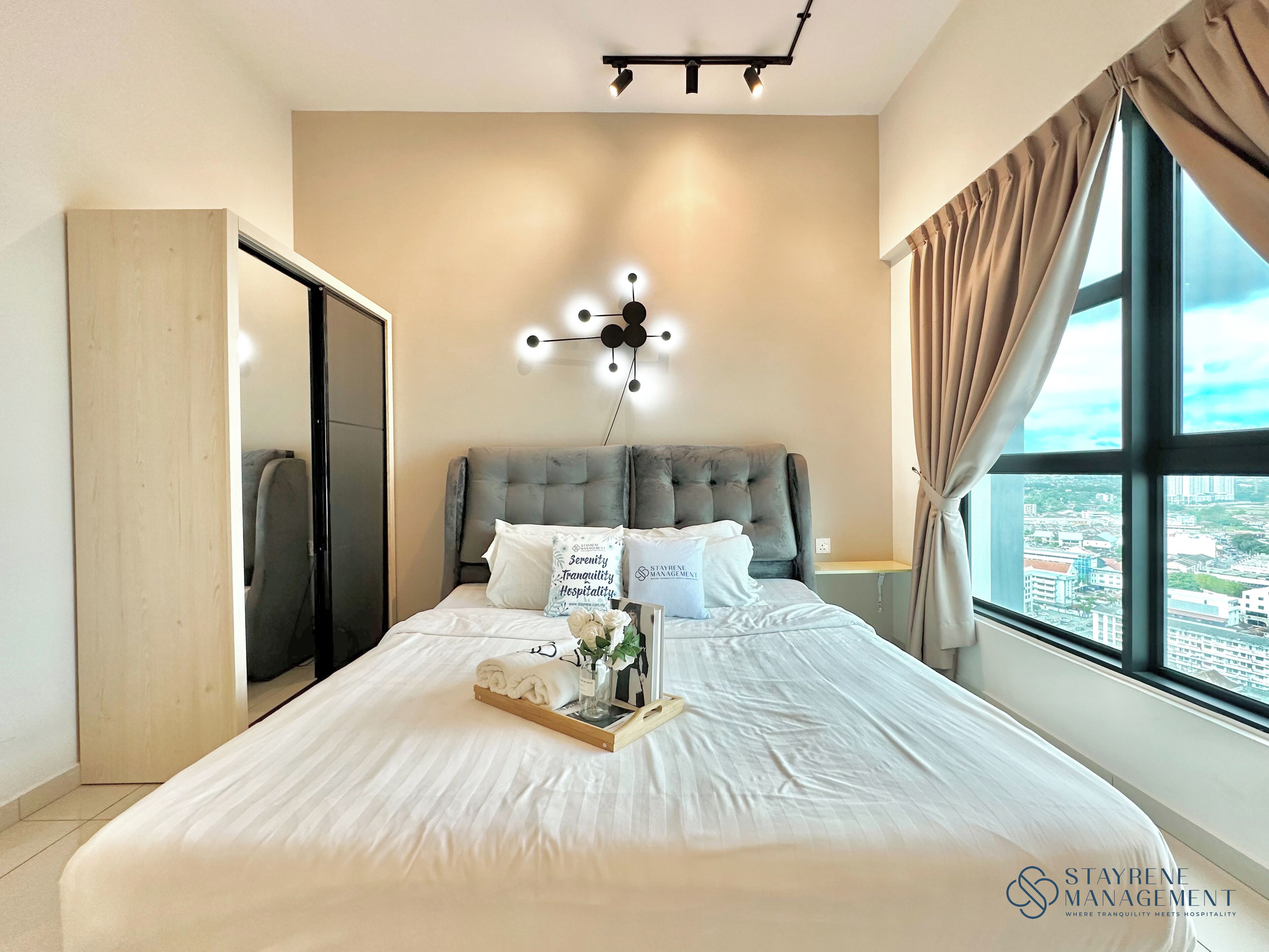 Quarto de hotel com uma cama grande, cabeceira, mesa de cabeceira, cortinas e vista para a cidade.