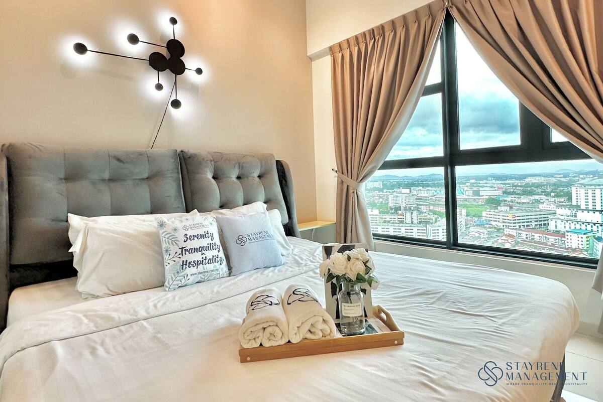 A bedroom with a large bed, a city view, a modern light fixture, and a tray with towels and flowers.