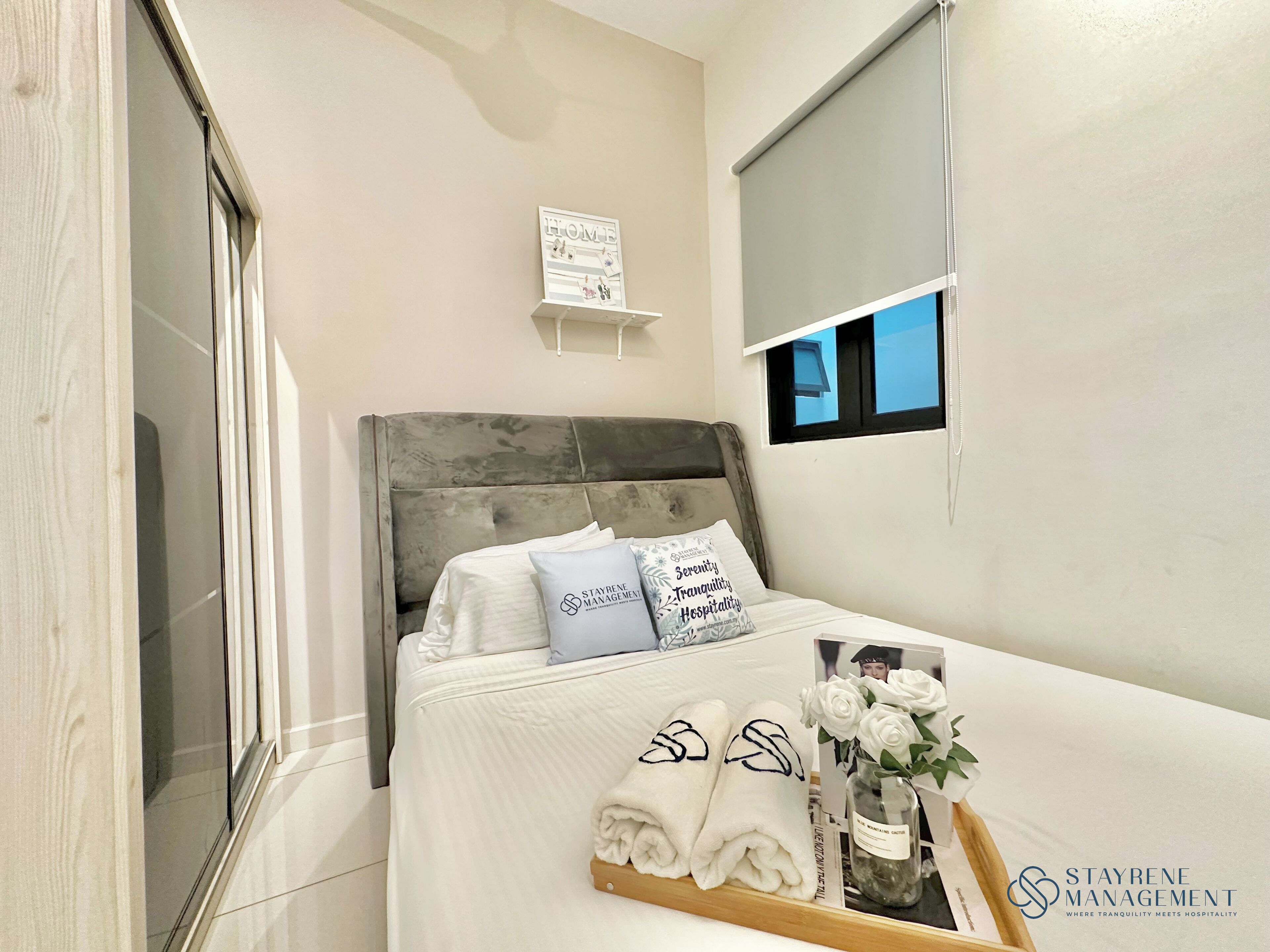 A hotel room with a bed, a bedside table with a tray of towels, a vase with flowers, and a sign on the wall.