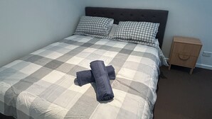 Ein ordentlich bezogenes Bett mit kariertem Überwurf und zwei Kissen, ein Nachttisch aus Holz und ein an der Wand befestigter Projektor.