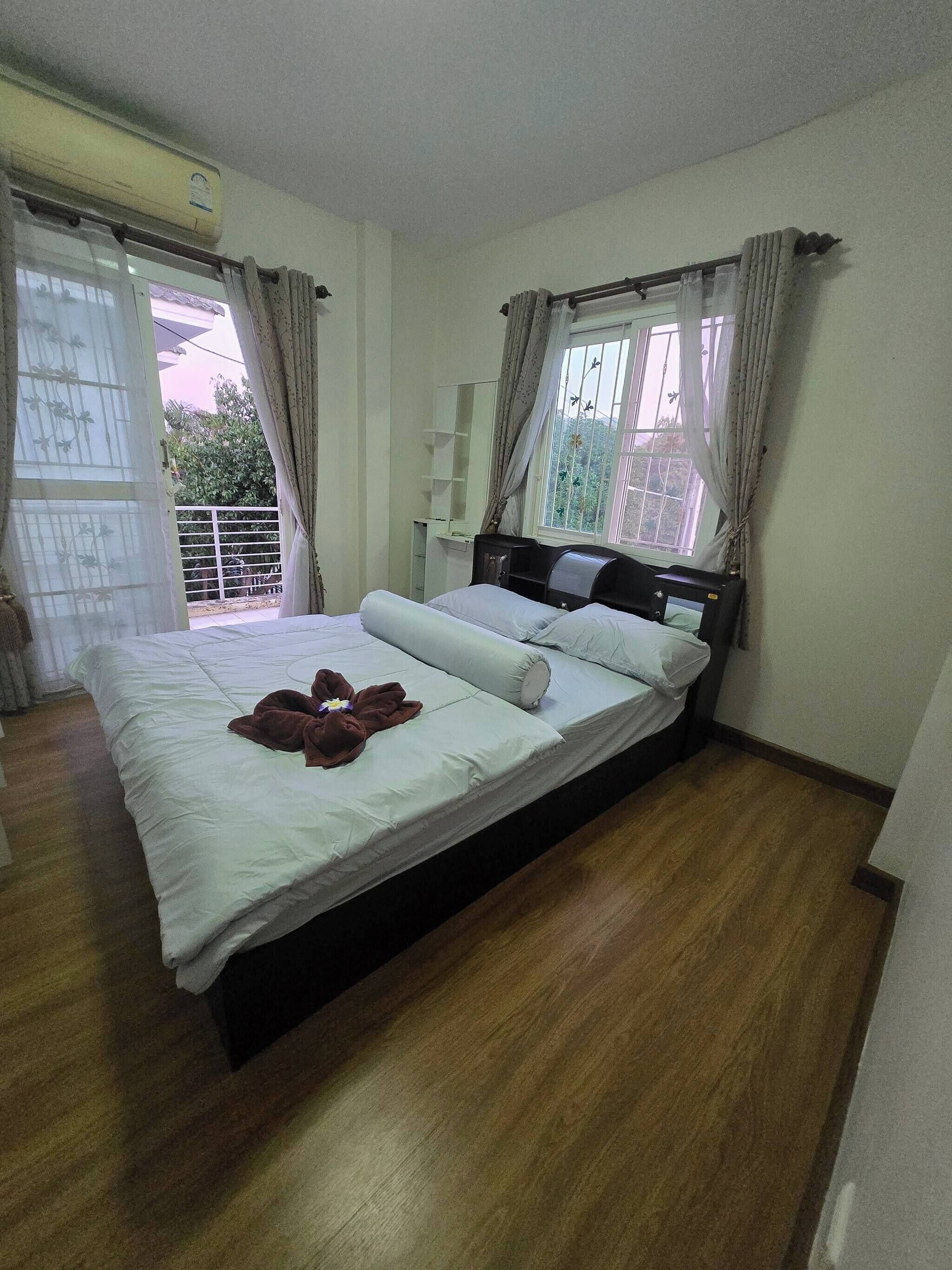 A bedroom with a wooden bed, a nightstand, a window with curtains, and a view of greenery outside.