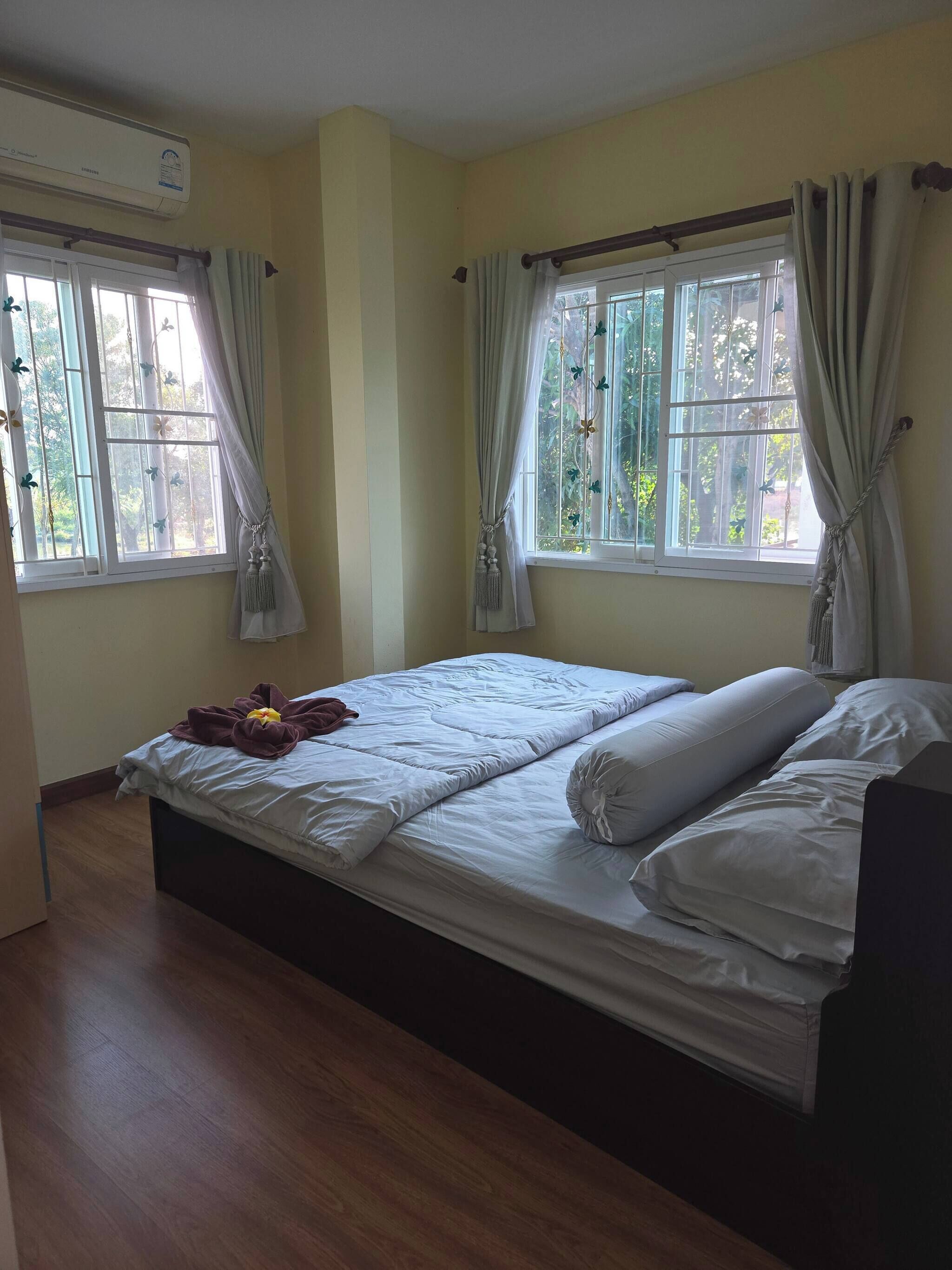 Quarto de hotel com uma cama, duas janelas com cortinas, um ar condicionado e piso de madeira.