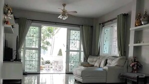 A spacious living room with a white sofa, a ceiling fan, and large windows with curtains. - Charming 3-bedroom House With Pool Near Lat Krabang Airport (Bangkok)