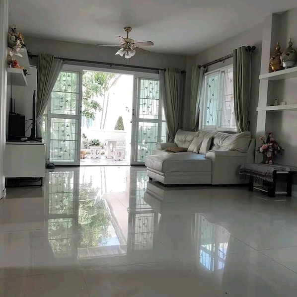 Un salon spacieux avec un canapé blanc, un ventilateur de plafond et de grandes fenêtres dotées de rideaux.