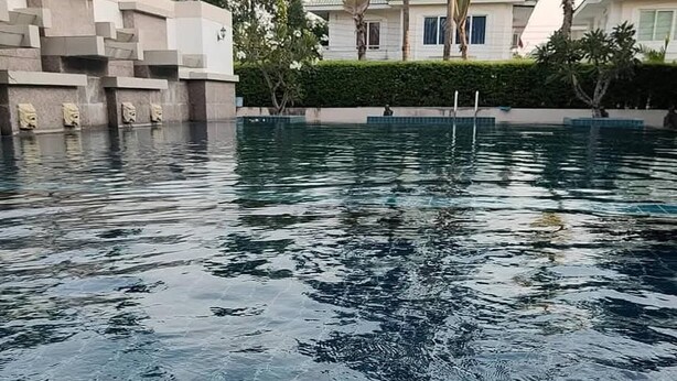 A swimming pool with clear water, surrounded by a landscaped area with palm trees and a building in the background. - Charming 3-bedroom House With Pool Near Lat Krabang Airport (Bangkok)