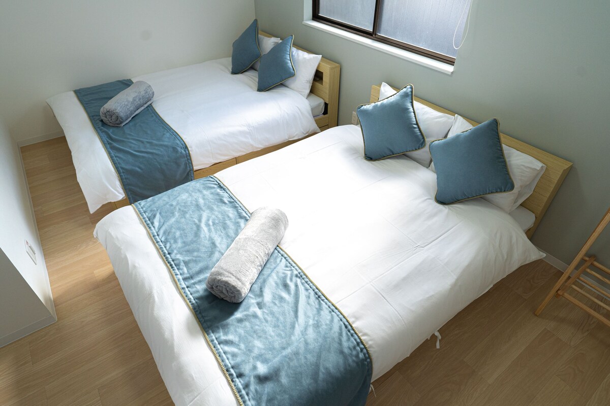 Two twin beds with blue bedding and pillows, a wooden floor, and a window with blinds.