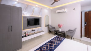 A modern hotel room with a flat-screen TV, a ceiling fan, an air conditioning unit, a desk with chairs, and a decorative rug. - Hotel Maharani Palace (varanasi)