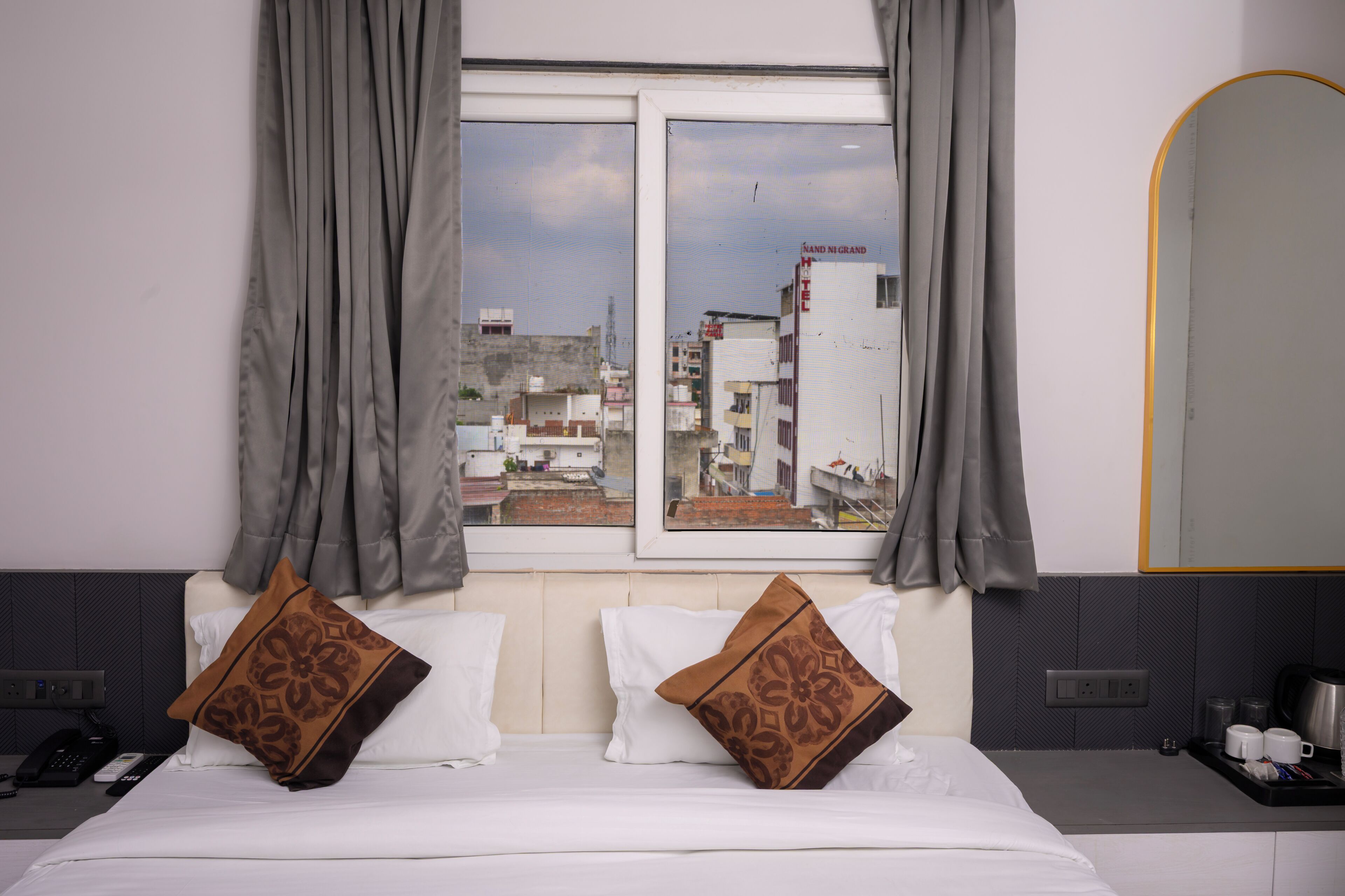 Une chambre d’hôtel avec un lit, deux oreillers, une fenêtre avec des rideaux, un miroir et une vue sur le paysage urbain.