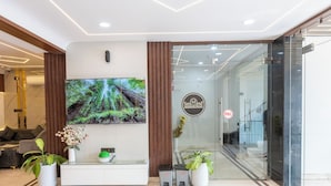 A reception area with a large flat-screen TV mounted on the wall, a reception desk with a white surface, and a glass door leading to the interior. - Hotel Maharani Palace (varanasi)