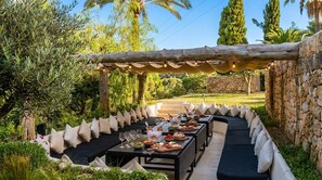 An outdoor dining area with a long table, cushioned seating, and a pergola overhead. - Villa Can Nikita – Amazing Experience in Ibiza – Relax in Nature and Luxury (Ibiza)