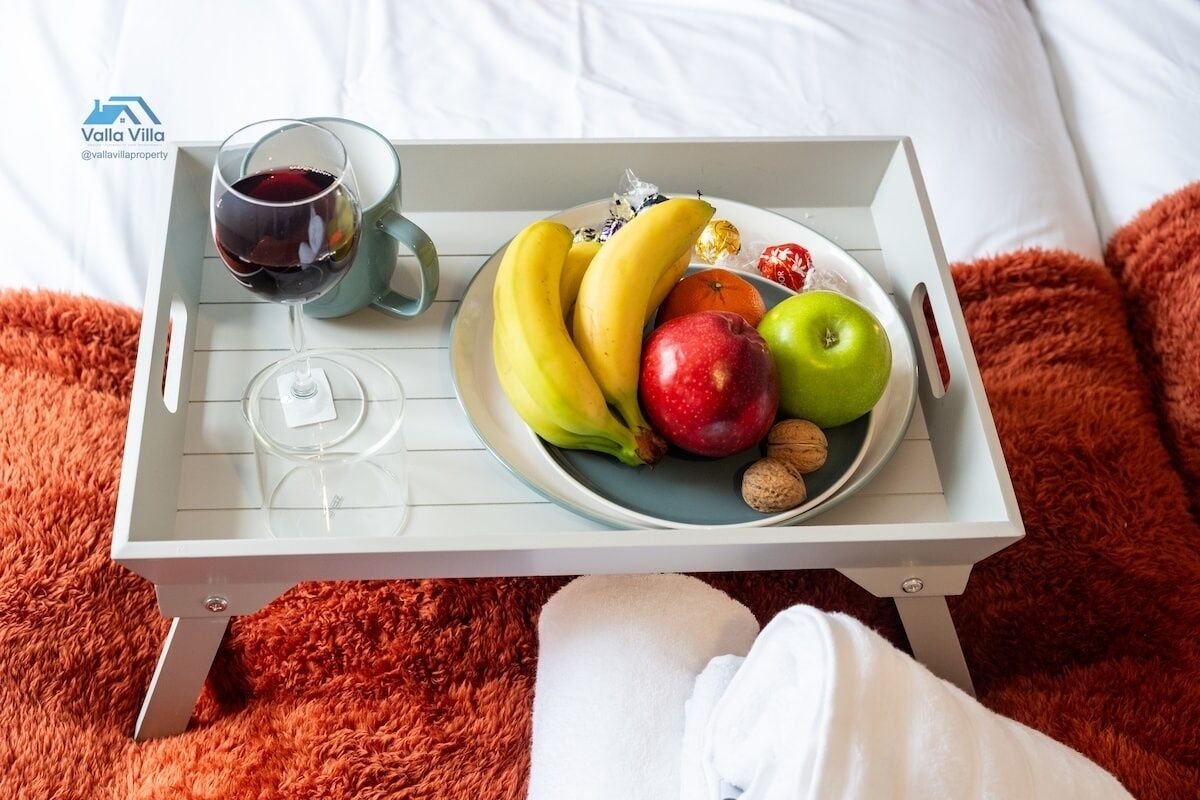 A tray with fruits, nuts, a glass of red wine, and a mug on a bed with a fluffy orange blanket.