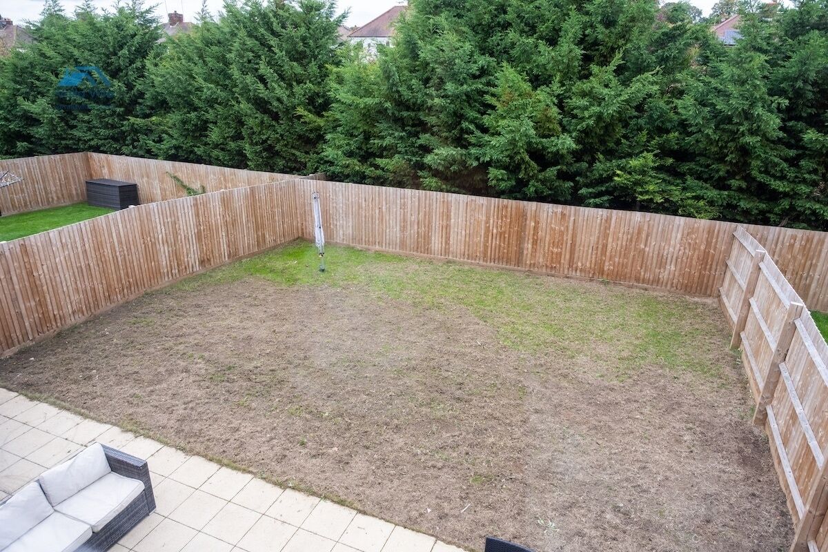 A backyard with a wooden fence, a small grassy area, and a tiled patio with a white sofa.