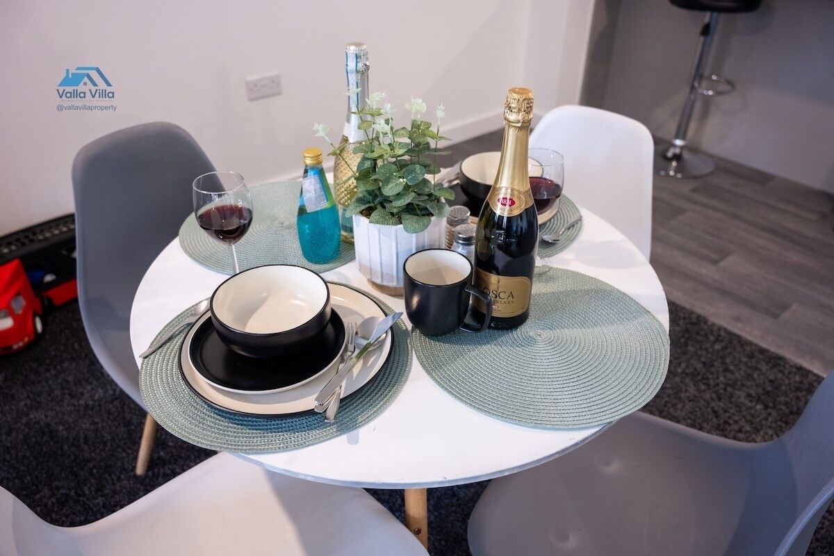 A dining table set with plates, cutlery, a bottle of champagne, a glass of red wine, a blue bottle, and a small potted plant.