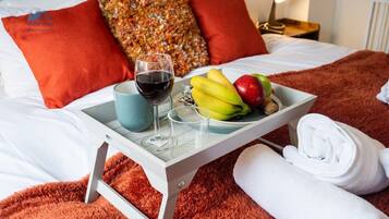 A neatly made bed with a tray of breakfast items including bananas, an apple, and a glass of red wine.