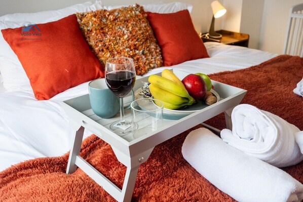 A neatly made bed with a tray of breakfast items including bananas, an apple, and a glass of red wine.