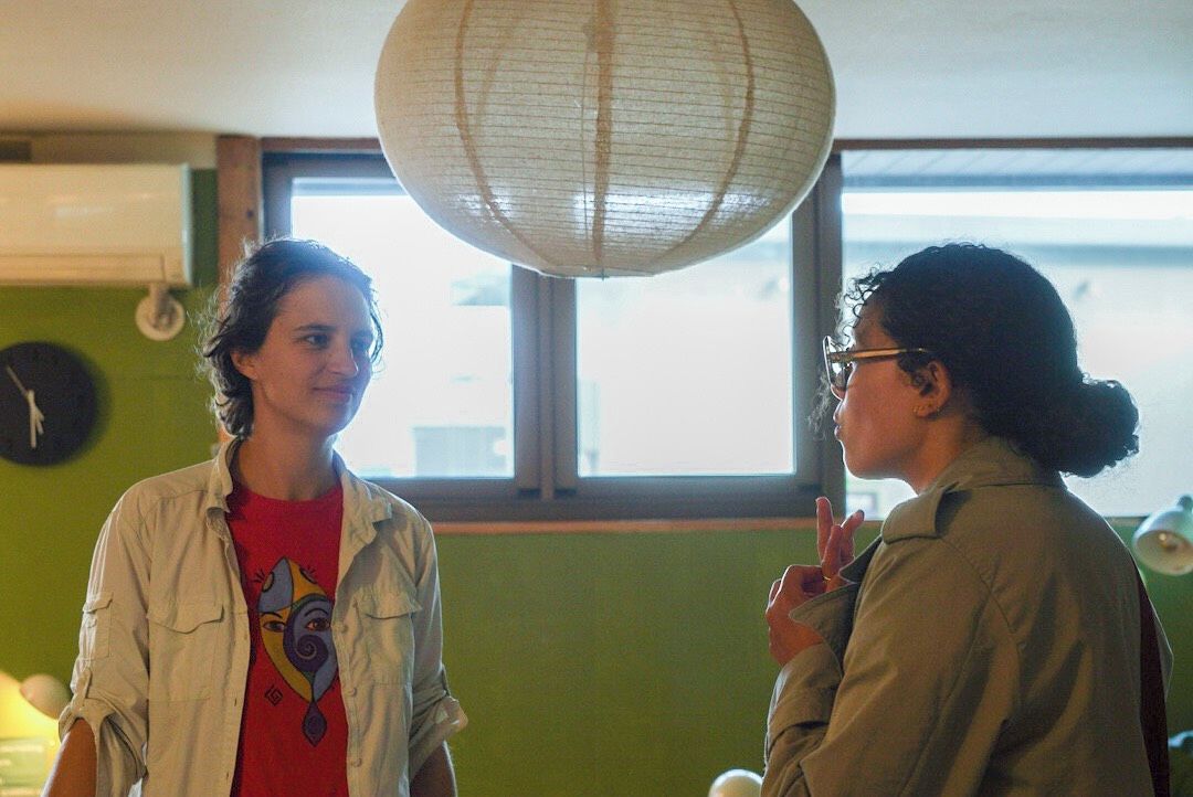 Two individuals in a room with a large window, a wall clock, and a hanging paper lantern.