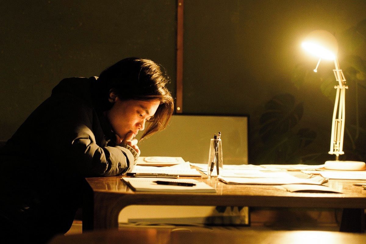 A person sitting at a desk with a desk lamp, papers, and a pen.