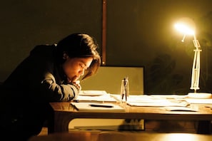 A person sitting at a desk with a desk lamp, papers, and a pen.