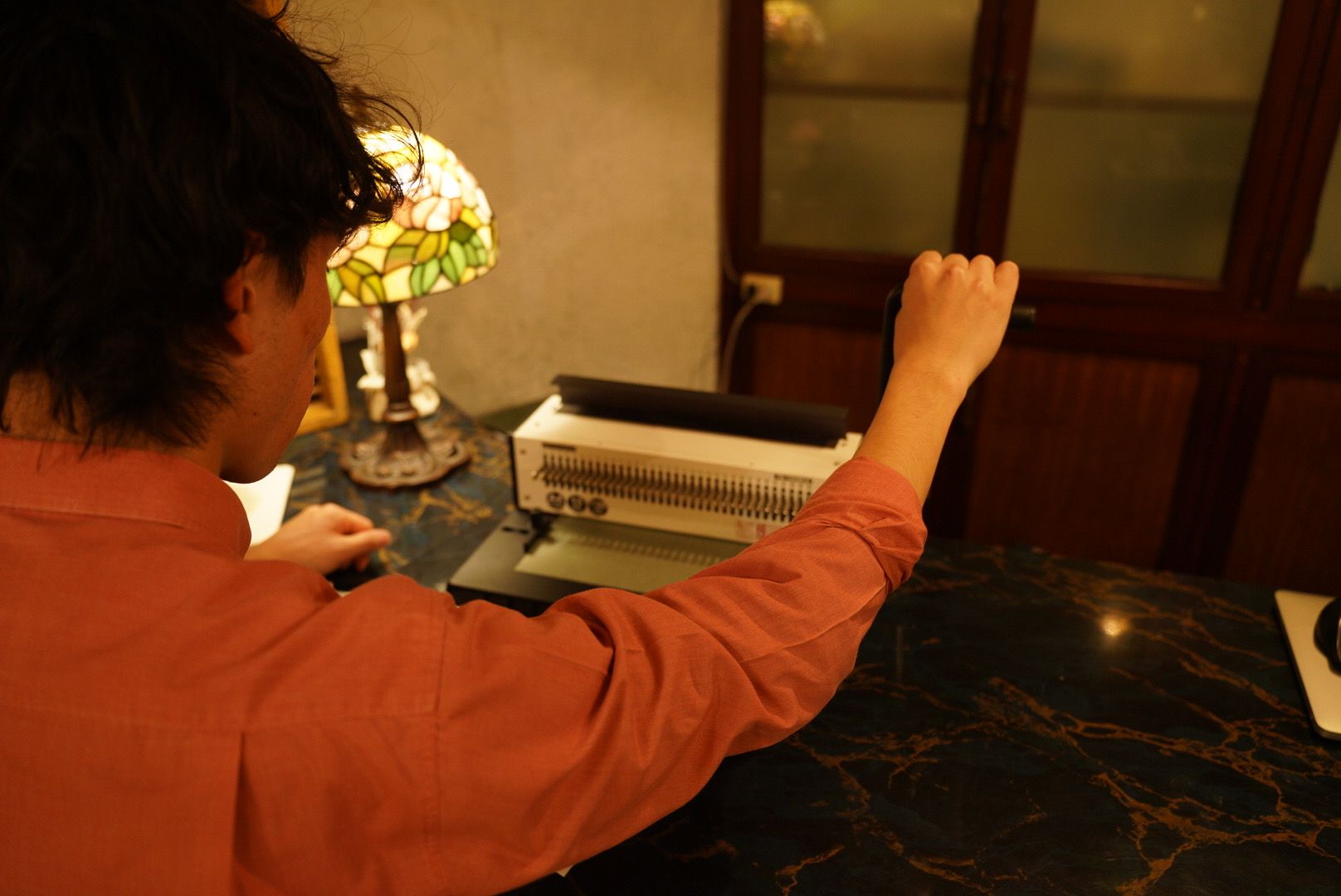 Una persona sentada a una mesa con una máquina de escribir, una lámpara y una ventana al fondo.