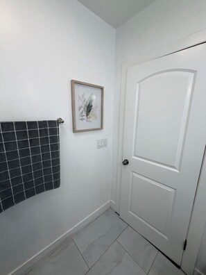 Un baño con una puerta blanca, un cuadro en la pared y una toalla colgada en un perchero.