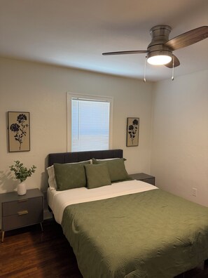 Un dormitorio con una cama, mesitas de noche, un ventilador de techo y una obra de arte en la pared.