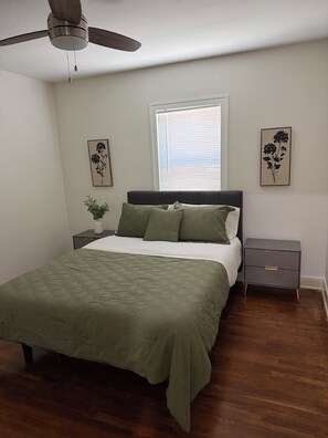 A bedroom with a bed, a nightstand, a bedside table, a ceiling fan, a window with blinds, and two wall-mounted pictures.