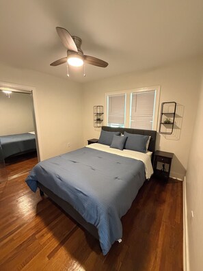 A bedroom with a bed, a ceiling fan, a window with blinds, and a nightstand with a lamp.