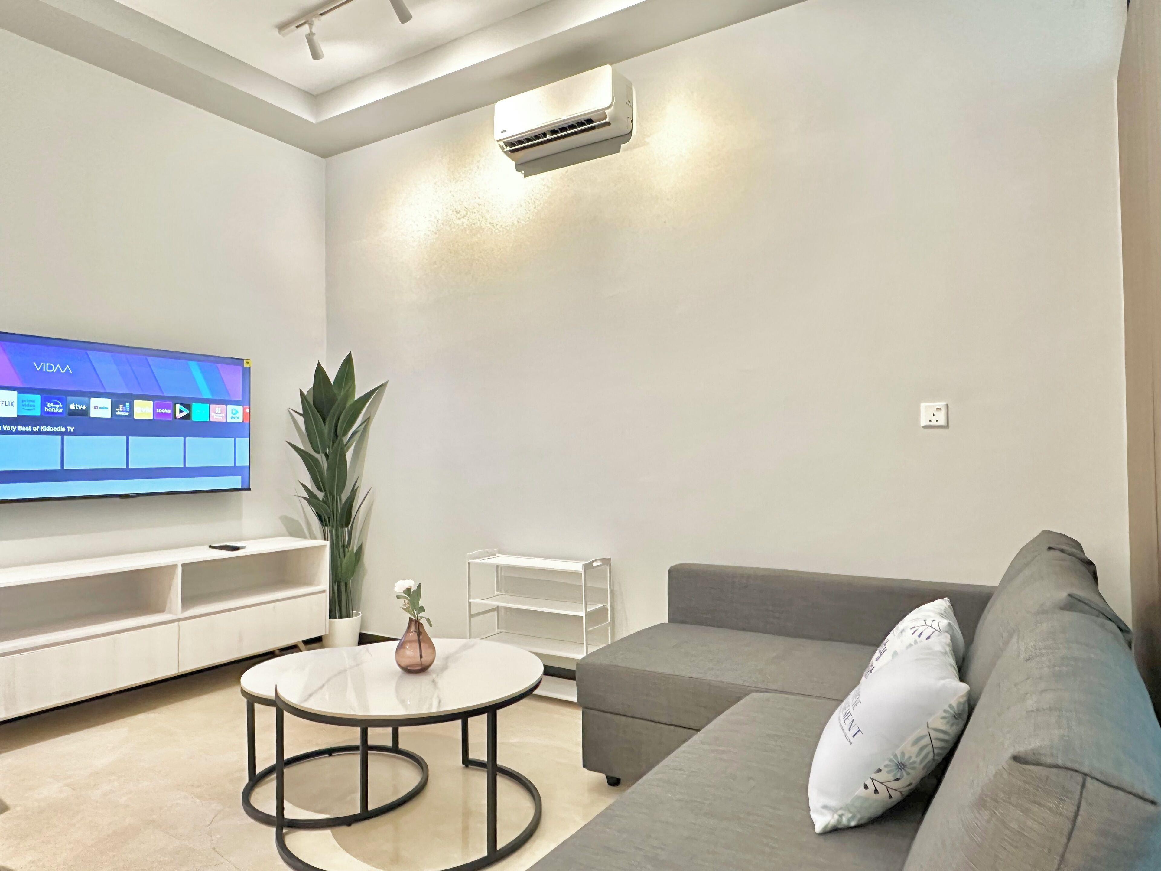 Een moderne woonkamer met een flatscreen televisie, een grijze bank, een ronde salontafel en een potplant.